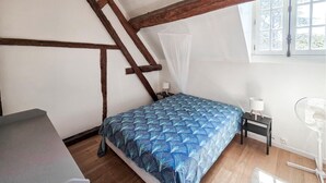 A bedroom with a sloped ceiling, a bed with a patterned bedspread, a nightstand with a lamp, a standing fan, and a window with a view of trees. - Nice home in Manthelan (Manthelan)