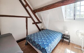 A bedroom with a sloped ceiling, a bed with a patterned bedspread, a nightstand with a lamp, a standing fan, and a window with a view of trees. - Nice home in Manthelan (Manthelan)