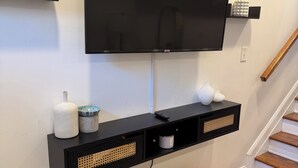 Televisão de tela plana fixada na parede com um móvel preto embaixo, dois pufes de macramé amarelo e branco, e uma escada à direita.