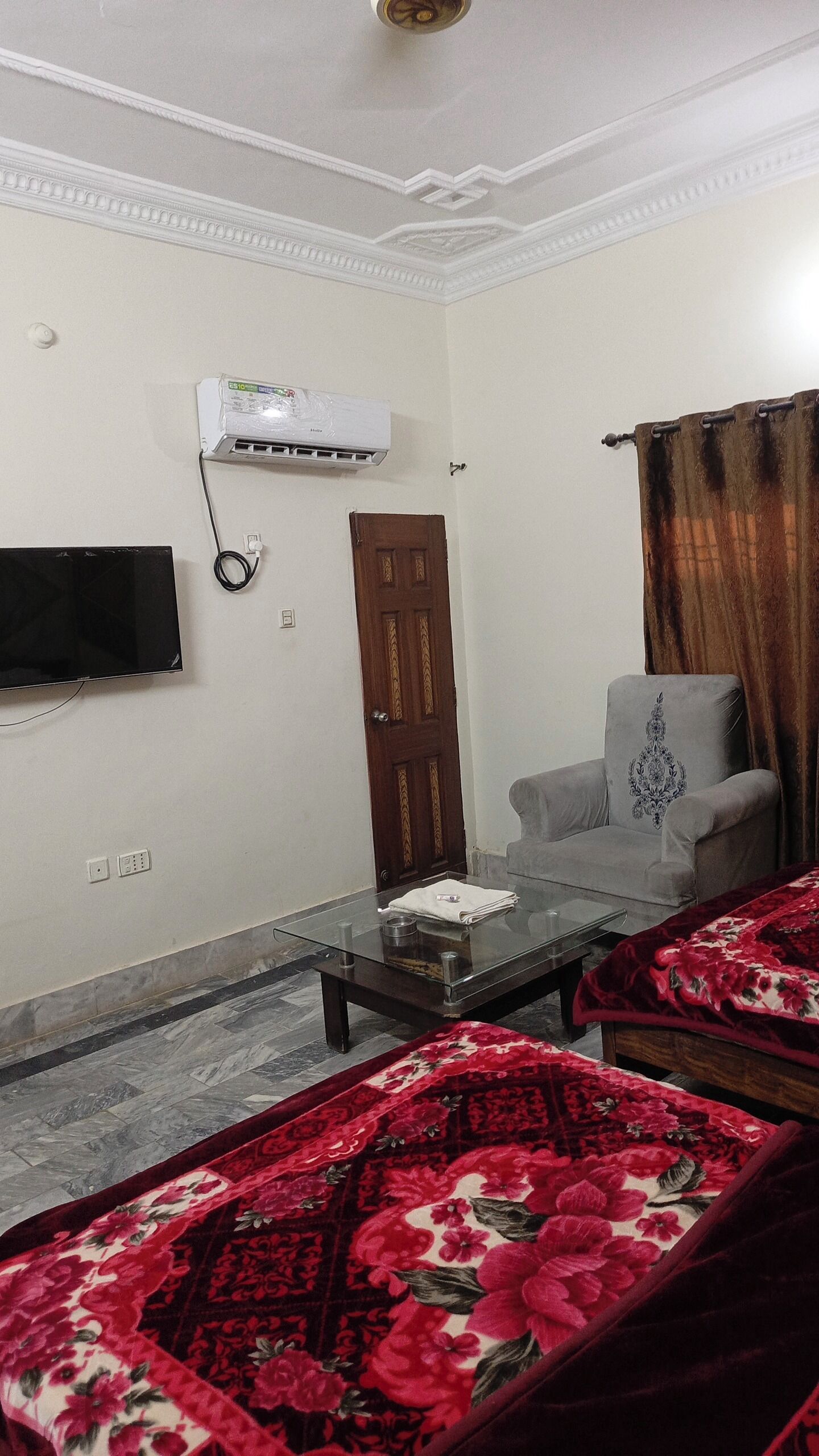 A living room with a flat-screen TV, a sofa, a coffee table, a patterned rug, a chair, an air conditioning unit, and a door.