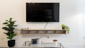 Una televisione a schermo piatto montata su una parete, sopra un ripiano in legno con un telecomando, libri e una candela.
