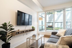 Ein Wohnzimmer mit Fernseher an der Wand, einer Couch, einem Couchtisch und einem großen Fenster mit Blick ins Freie.