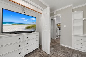 A room with a television on a white dresser, a wooden herringbone floor, and a white door leading to another room. - Ocean Springs Retreat, 5 min to downtown, Sleeps 8 (Ocean Springs)
