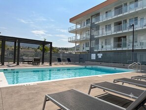 A swimming pool with lounge chairs, a covered seating area, and a multi-story building with balconies.
