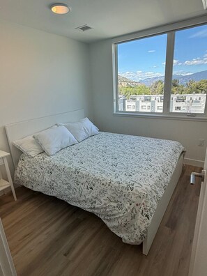 Une chambre avec une grande fenêtre, un lit recouvert d’une couvre-lit à motifs floraux et une tête de lit blanche.