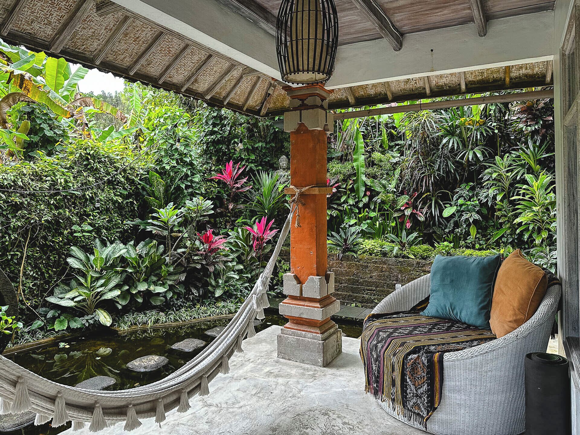 Un sillón hamaca con cojines, un pilar de piedra y abundante vegetación.
