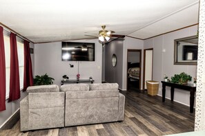 Ein Wohnzimmer mit grauem Sofa, Deckenventilator, Flachbildfernseher, kleinem Tisch mit Pflanzen und einem Korbwäscher.