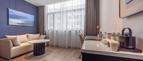 Habitación de hotel moderna con sofá, una pequeña mesa redonda, un escritorio con silla, cafetera y vistas a la ciudad a través de amplias ventanas.