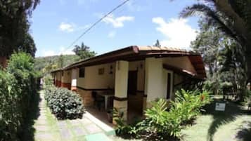 Uma casa térrea com varanda coberta, cercada por vegetação e um céu límpido.