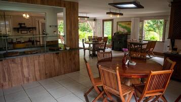 A spacious dining area with a wooden table and chairs, a kitchen with a glass counter, and large windows offering a view of greenery.