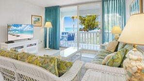 Habitación de hotel con televisión, muebles de mimbre blanco, una mesa de centro de vidrio y vista a la playa.