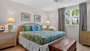 Quarto com cama, duas mesas de cabeceira, um criado-mudo, um banco de madeira, janela com cortinas e duas luminárias de parede.