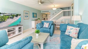 A living room with teal sofas, a coffee table, a TV, and a staircase. - Harbour Heights #26 Seven Mile Beach, 2 Bedroom, 2.5 Bathroom Second Floor Townhouse (Seven Mile Beach)