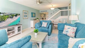 A living room with teal sofas, a coffee table, a TV, and a staircase. - Harbour Heights #26 Seven Mile Beach, 2 Bedroom, 2.5 Bathroom Second Floor Townhouse (Seven Mile Beach)