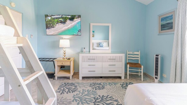 A bedroom with a bed, a bedside table with a lamp, a dresser, a chair, a painting of a beach, and a radiator. - Harbour Heights #26 Seven Mile Beach, 2 Bedroom, 2.5 Bathroom Second Floor Townhouse (Seven Mile Beach)