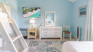 A bedroom with a bed, a bedside table with a lamp, a dresser, a chair, a painting of a beach, and a radiator. - Harbour Heights #26 Seven Mile Beach, 2 Bedroom, 2.5 Bathroom Second Floor Townhouse (Seven Mile Beach)