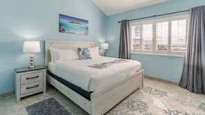 A bedroom with a bed, nightstands, a lamp, a painting, a window with curtains, and a patterned rug. - Harbour Heights #26 Seven Mile Beach, 2 Bedroom, 2.5 Bathroom Second Floor Townhouse (Seven Mile Beach)