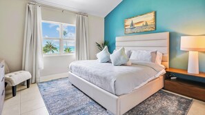 A bedroom with a bed, a nightstand, a lamp, a painting, and a view of the ocean through a window. - Harbour Heights #38 Seven Mile Beach, 2 Bed, 2 Bath Second Floor Townhouse (Seven Mile Beach)