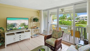 A balcony with a TV, wicker furniture, and a view of a pool and greenery. - Harbour Heights #8 Seven Mile Beach, 2 Bed, 2 Bath Second Floor Townhouse (Seven Mile Beach)