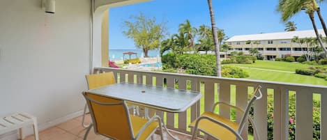 A balcony with a table and chairs, overlooking a pool and a building with a white roof.