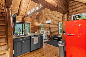 Interior de una cabaña de troncos con una zona de cocina, paredes de madera y un refrigerador rojo.
