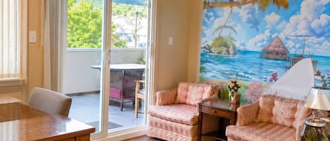 A room with a wooden dining table, two armchairs, a sofa, and a view of a tropical beach through a large window.