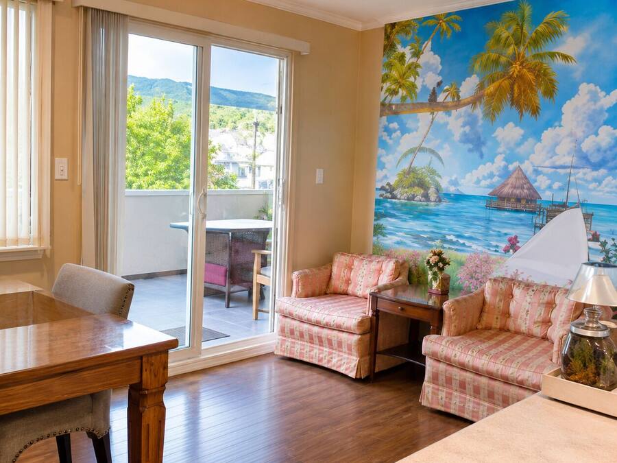Une pièce comprenant une table à manger en bois, deux fauteuils, un canapé et une vue sur une plage tropicale, visible à travers une grande fenêtre.