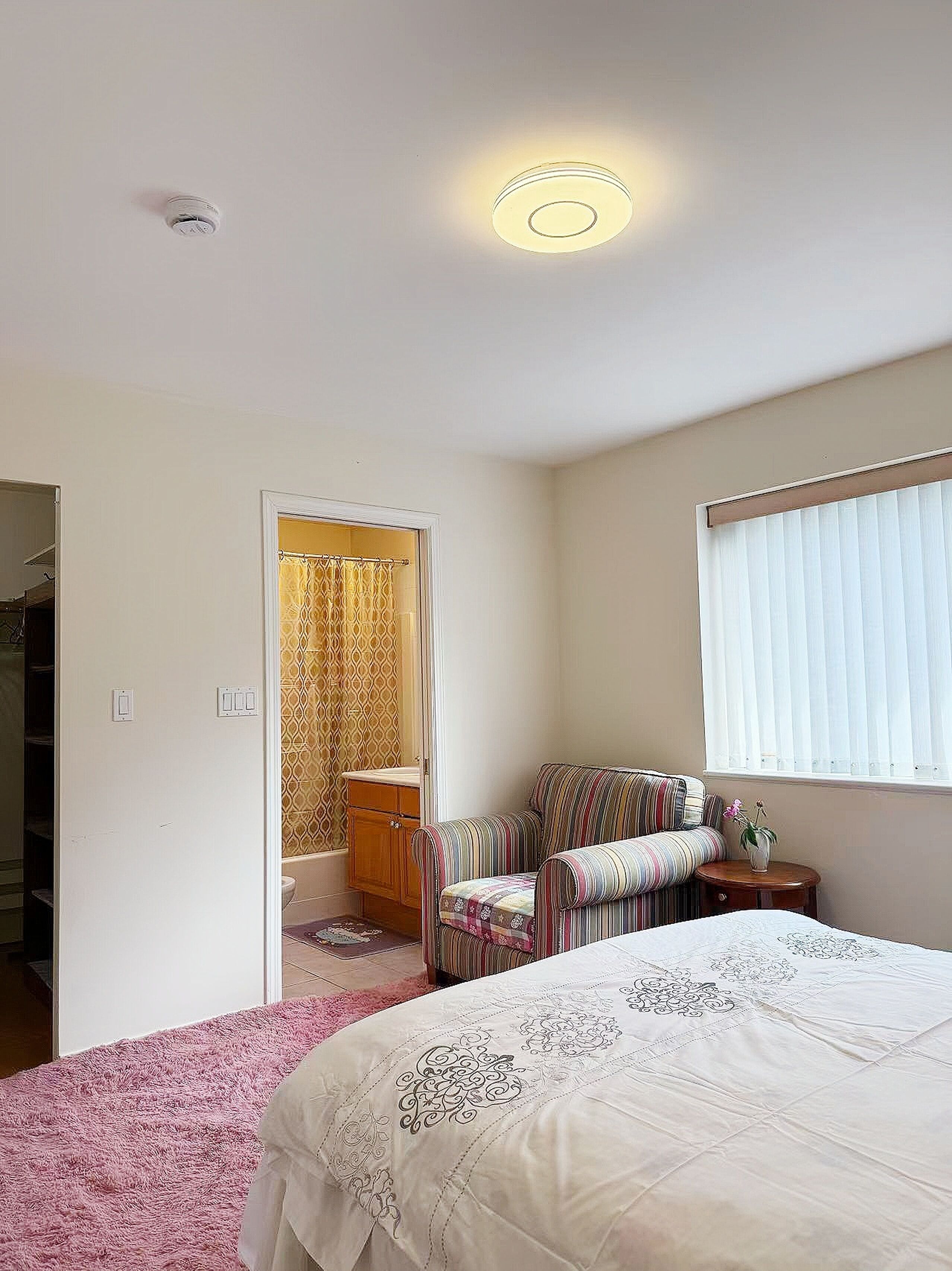 Un dormitorio con cama, una silla, una mesita, un ventanal con persianas y una puerta que da al baño.