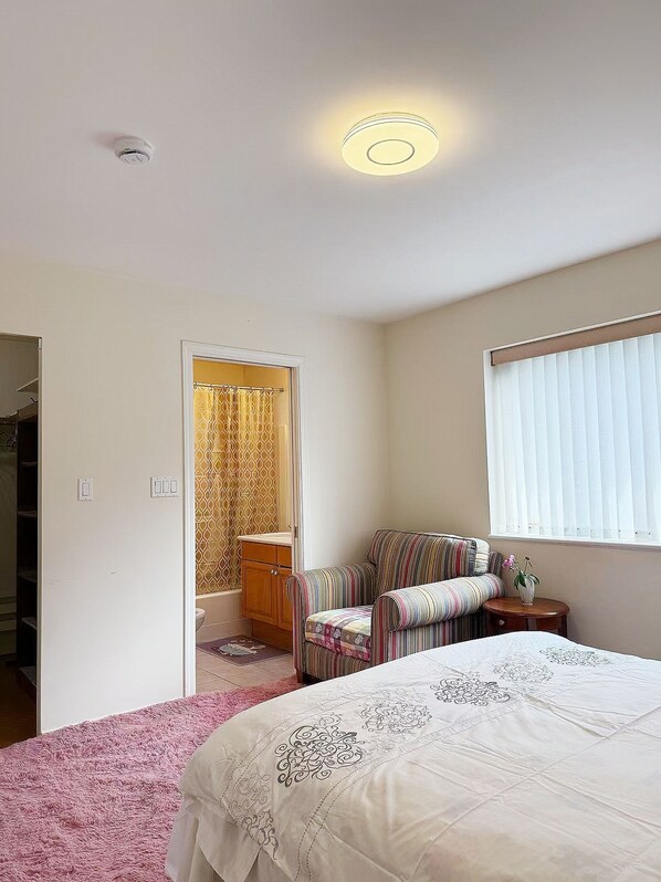 A bedroom with a bed, a chair, a small table, a window with blinds, and a door leading to a bathroom.