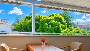 A balcony with a wooden table and two chairs, a vase with red flowers, and a view of green trees and houses. - Cozy Home in Vancouver (Vancouver)