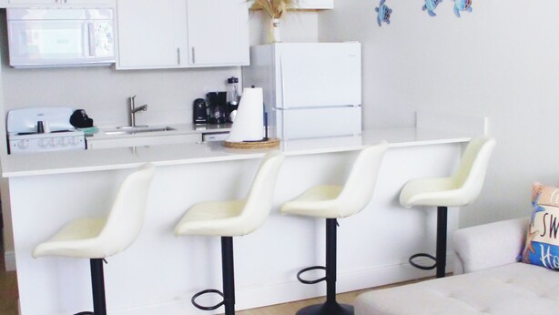 A modern kitchen with white cabinets, a microwave, a coffee maker, and a refrigerator. There are three bar stools with white seats and black bases. - Direct Oceanfront Daytona Beach Condo | Full Kitchen | Beach Access (Daytona Beach)