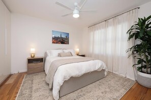 Un dormitorio con cama, dos mesitas de noche con lámparas, ventilador de techo, ventana grande con cortinas y un cuadro de paisaje urbano en la pared.