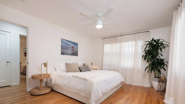 A bedroom with a bed, a ceiling fan, a window with curtains, a bedside table, and a potted plant.