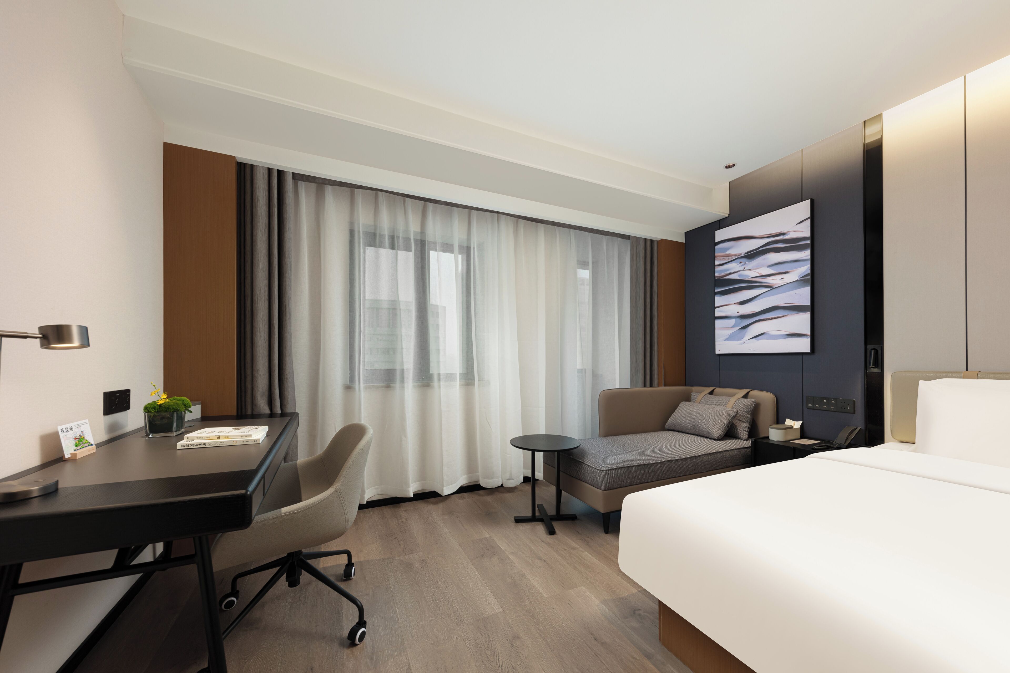Habitación de hotel con cama, escritorio, silla, mesita pequeña y un gran ventanal con cortinas.