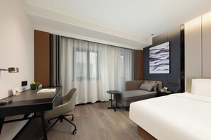 A hotel room with a bed, a desk, a chair, a small table, and a large window with curtains.