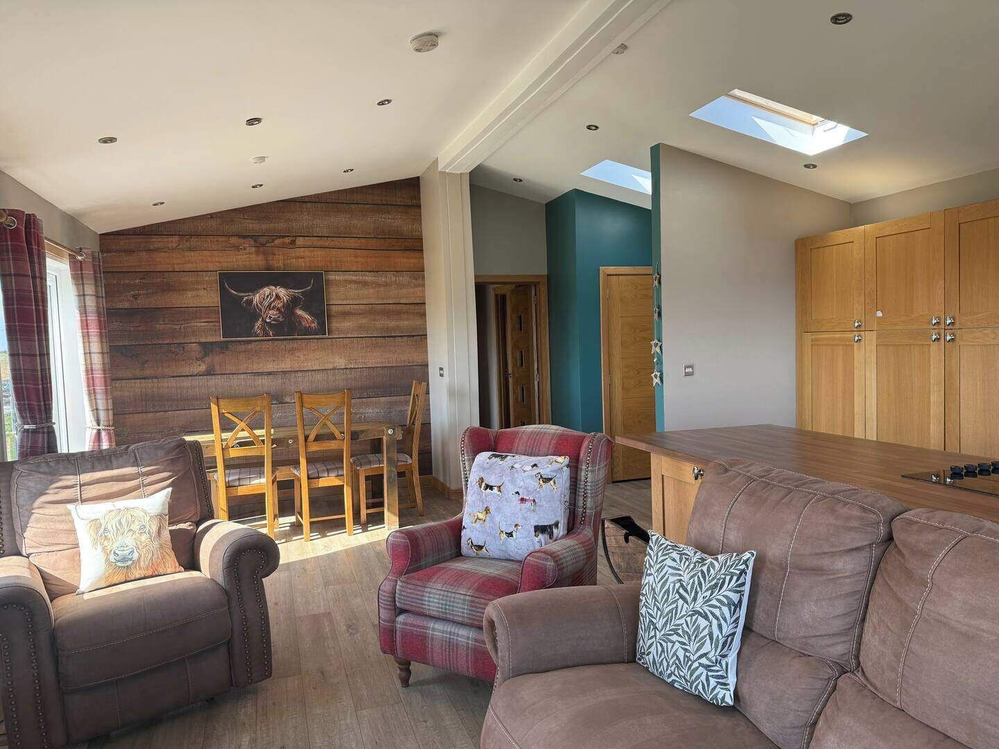 Un salon chaleureux avec un canapé, des fauteuils et une table à manger. Un mur en bois est décoré d’un tableau encadré, et une lucarne se trouve au-dessus de la table.