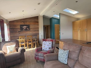 A cozy living room with a sofa, armchairs, and a dining table. There is a wooden wall with a framed picture and a skylight above the table.