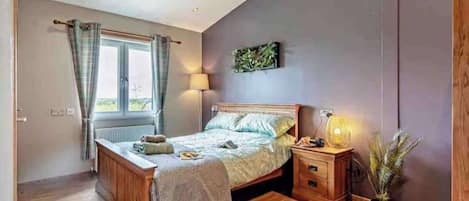 A bedroom with a wooden bed, bedside tables, a window with curtains, a wall-mounted lamp, and a potted plant.