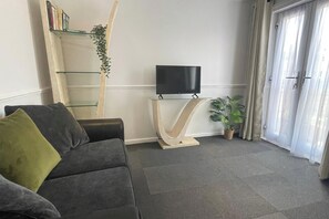 Ein Wohnzimmer mit einer Couch, einem Fernseher auf einem Schrank, einer Pflanze und einer mit Vorhängen versehenen Tür.