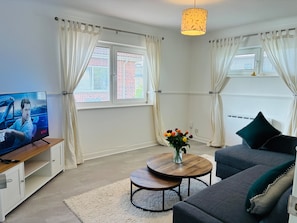 A modern living room with a television, a round coffee table, a sofa, and a decorative vase with flowers. - Cozy Grays Apartment (Grays)