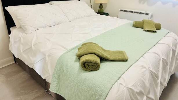 A neatly made bed with white linens and a green quilt, a lamp on a bedside table, and a rolled towel on the bed. - Cozy Grays Apartment (Grays)