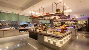 A modern hotel lobby with a central island featuring a buffet, surrounded by seating areas and a kitchen in the background.