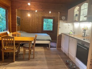 A cozy cabin interior with a wooden dining table, chairs, a bed, a small kitchen with a dishwasher, and a window with a view of greenery. - A cozy log cabin with a sauna and fireplace right next to the forest (Karwitz)