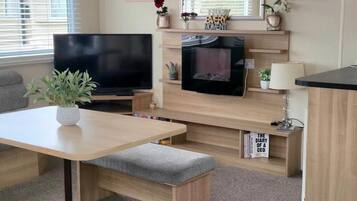 A modern living room with a flat-screen TV, a fireplace, a dining table with a bench, and a bookshelf with books.