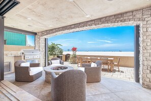 Eine Terrasse im Freien mit einer Steinsäule, einem Grill, Korbmöbeln und Blick auf den Strand.