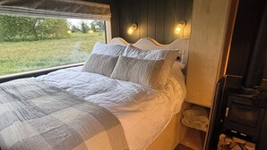 Una cama con una colcha a cuadros, una ventana con vistas a follaje y una lámpara fijada a la pared.