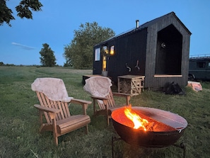 A rustic cabin with a fire pit, two covered chairs, and a small table.