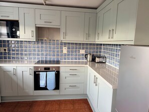 A kitchen with white cabinets, a tiled backsplash, a stove, a toaster, and a refrigerator. - SpaHome's By The Sea (Morpeth)