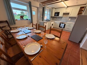 A dining area with a wooden table set for four, a kitchen with white cabinets and a microwave, and a view of the outdoors.