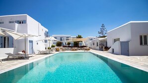 A clear blue pool surrounded by white buildings with balconies, a sun umbrella, and lounge chairs. - Aeolos Resort (Mykonos)